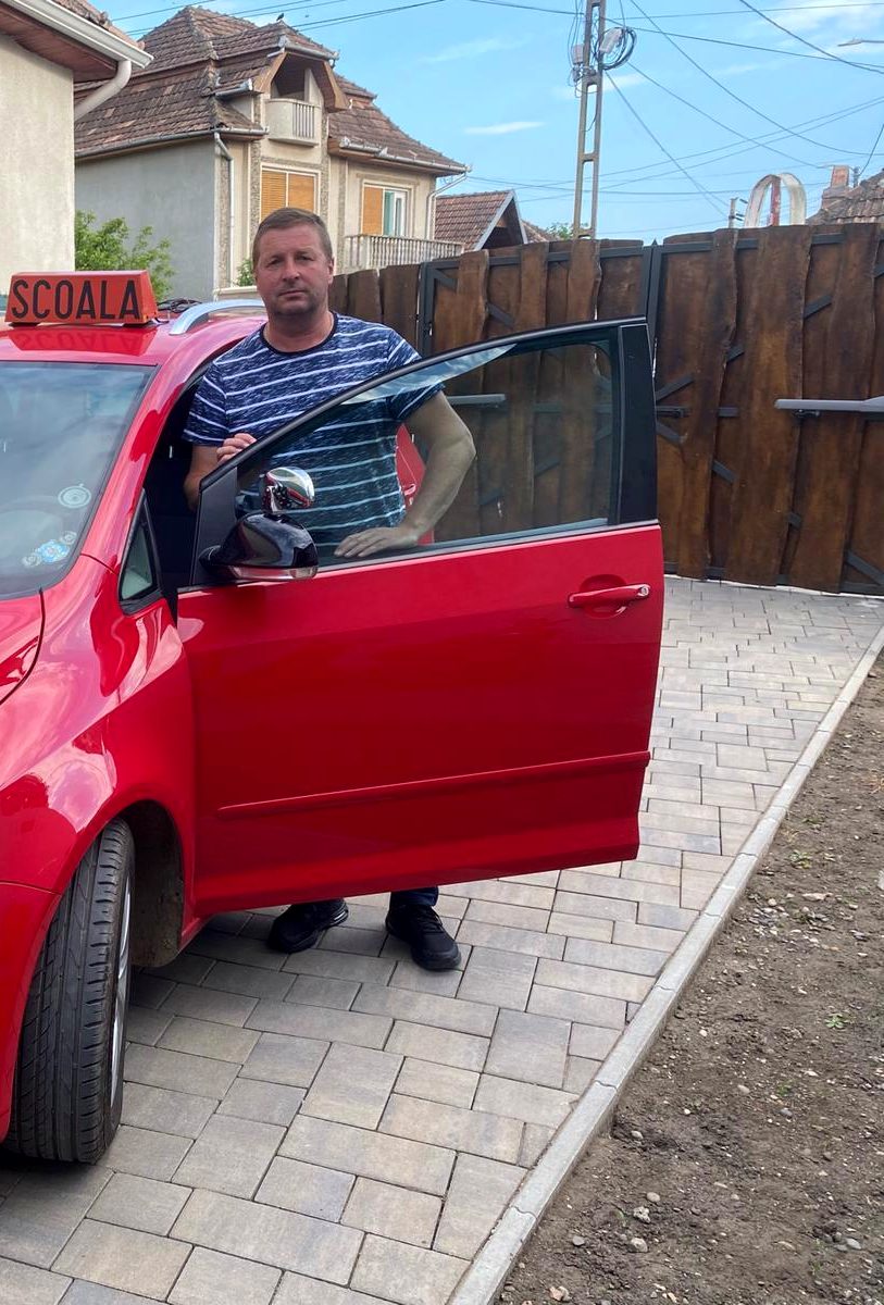 Mașina roșie a școlii AUTO LECHI, cu tăblița ȘCOALĂ pe plafon și instructorul la ușă